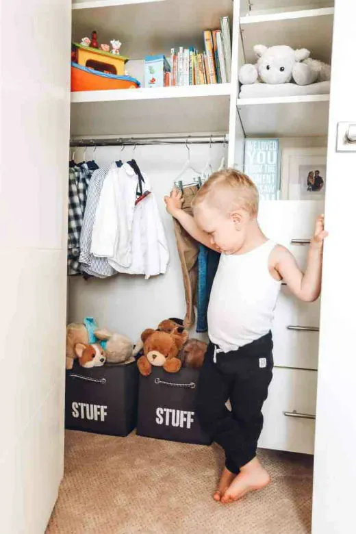 A toddler reaching clothes on a low rail in his built-in closet, storage bins labelled Stuff below