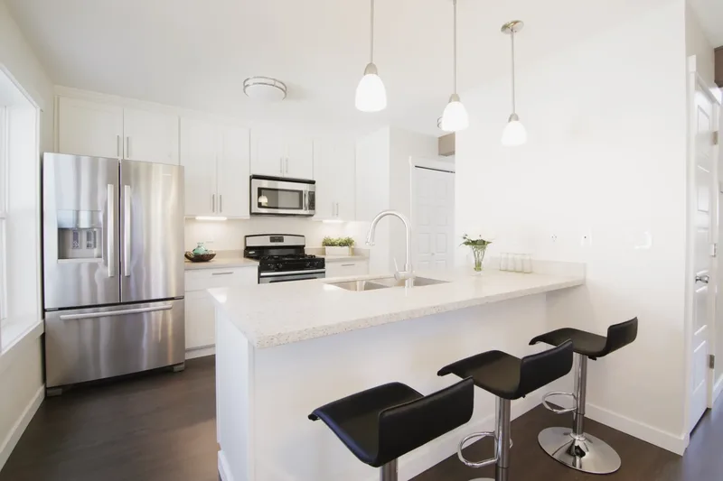 White shaker kitchen with a speckled peninsula and three black bar stools, stainless fridge against the far wall