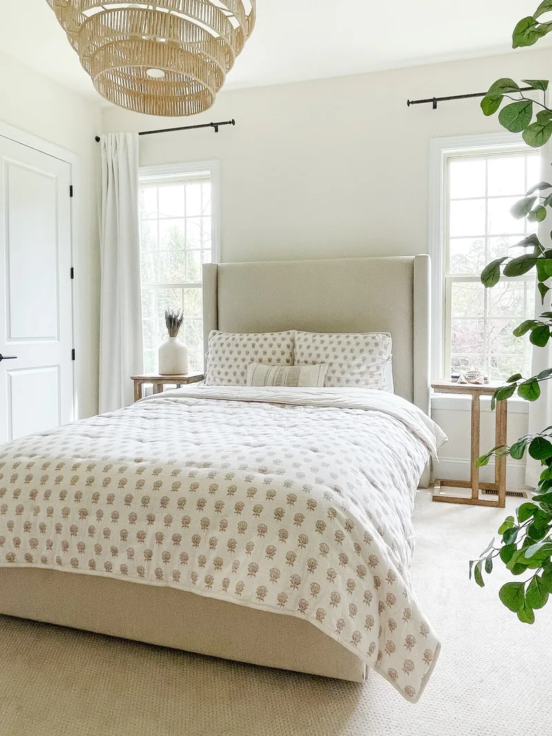 A linen wingback bed with a block-print quilt beneath a woven rattan chandelier, windows on both sides