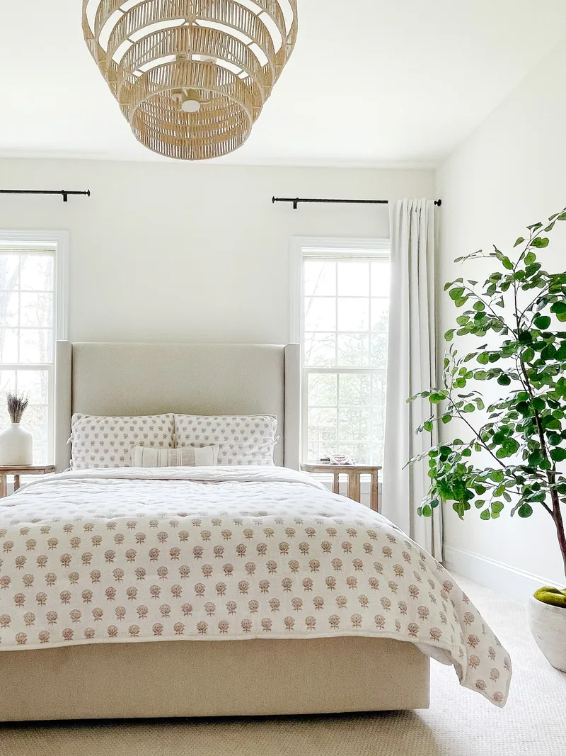 The bed from the foot of the room, a tall potted tree beside it under the rattan pendant