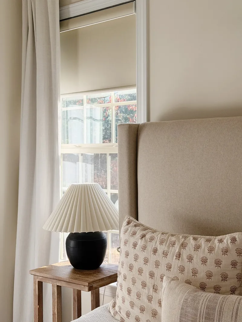 Close on the nightstand: a black lamp with pleated shade beside the linen headboard and printed pillows
