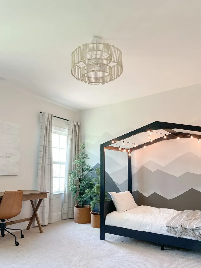 Dark navy house-frame bed strung with cafe lights against a painted mountain mural, desk and window beyond