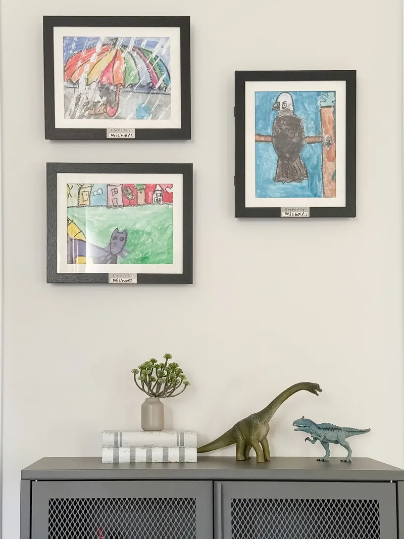 Close view of three framed children's paintings above a mesh cabinet, with toy dinosaurs and a potted succulent
