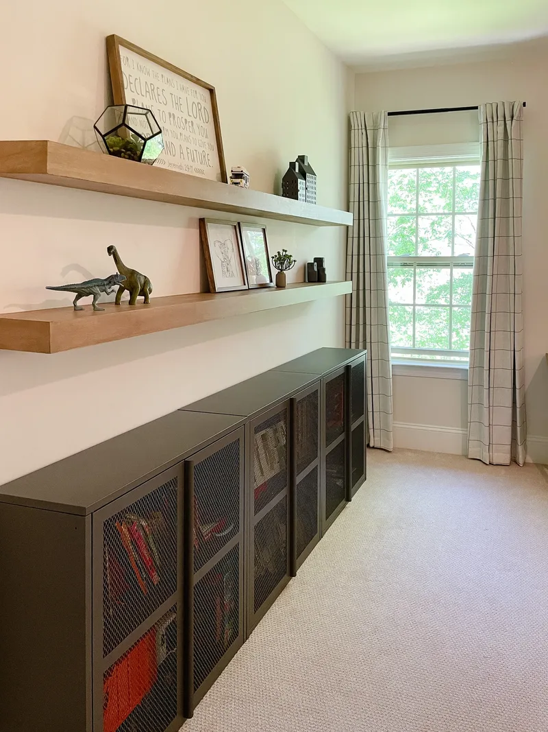Light oak floating shelves over low olive mesh-front cabinets, dinosaur figures above and check curtains at the window