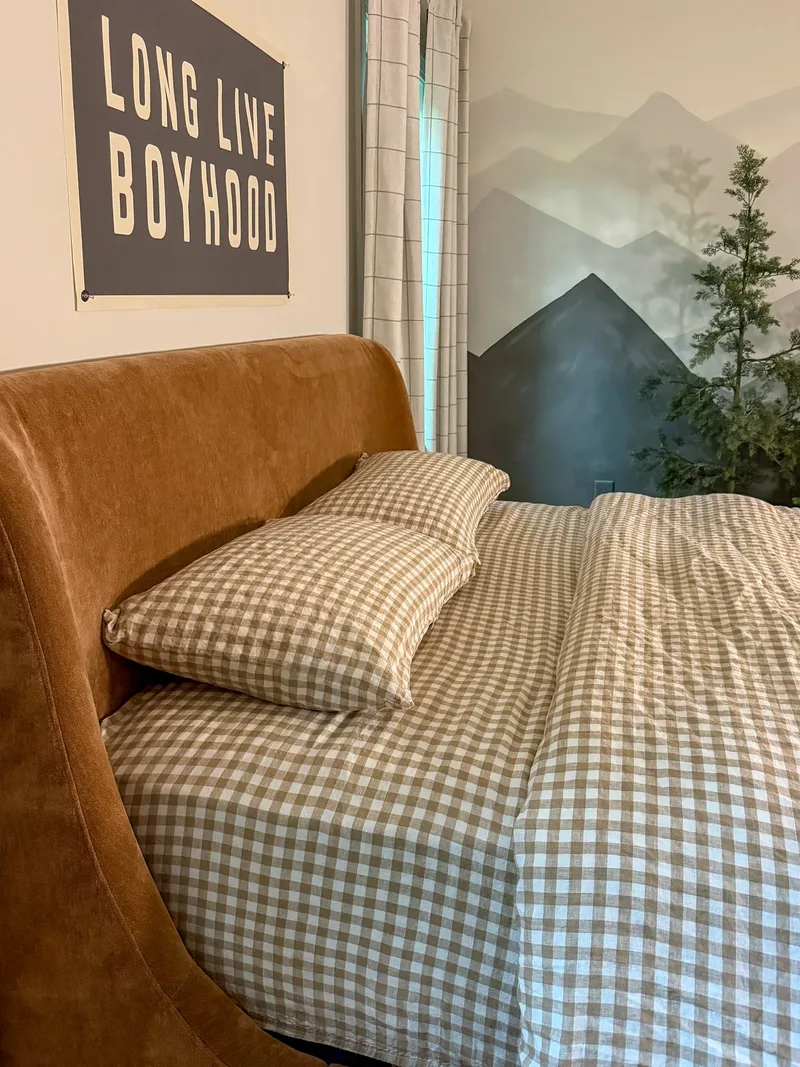 Close on the caramel suede headboard and tan gingham bedding, a Long Live Boyhood banner above