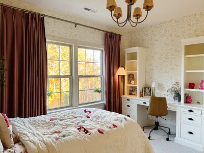 A white built-in desk between two bookcase towers, tan leather task chair, woodland wallpaper and rust velvet curtains