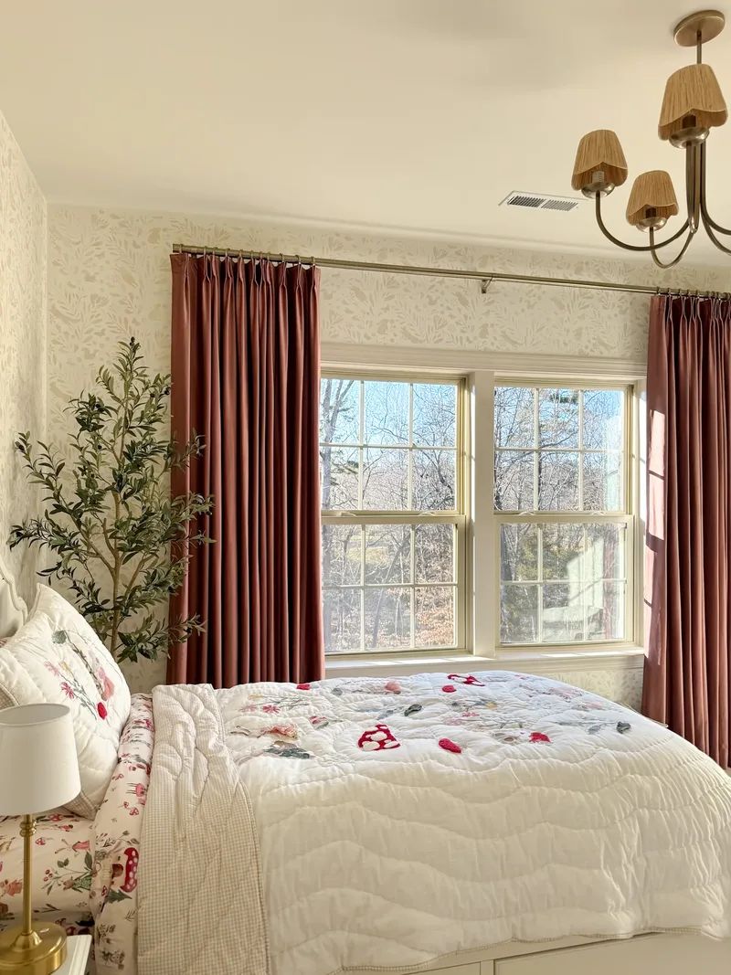 The bed from the other side, brass lamp and floral pillows in the foreground, olive tree by the window