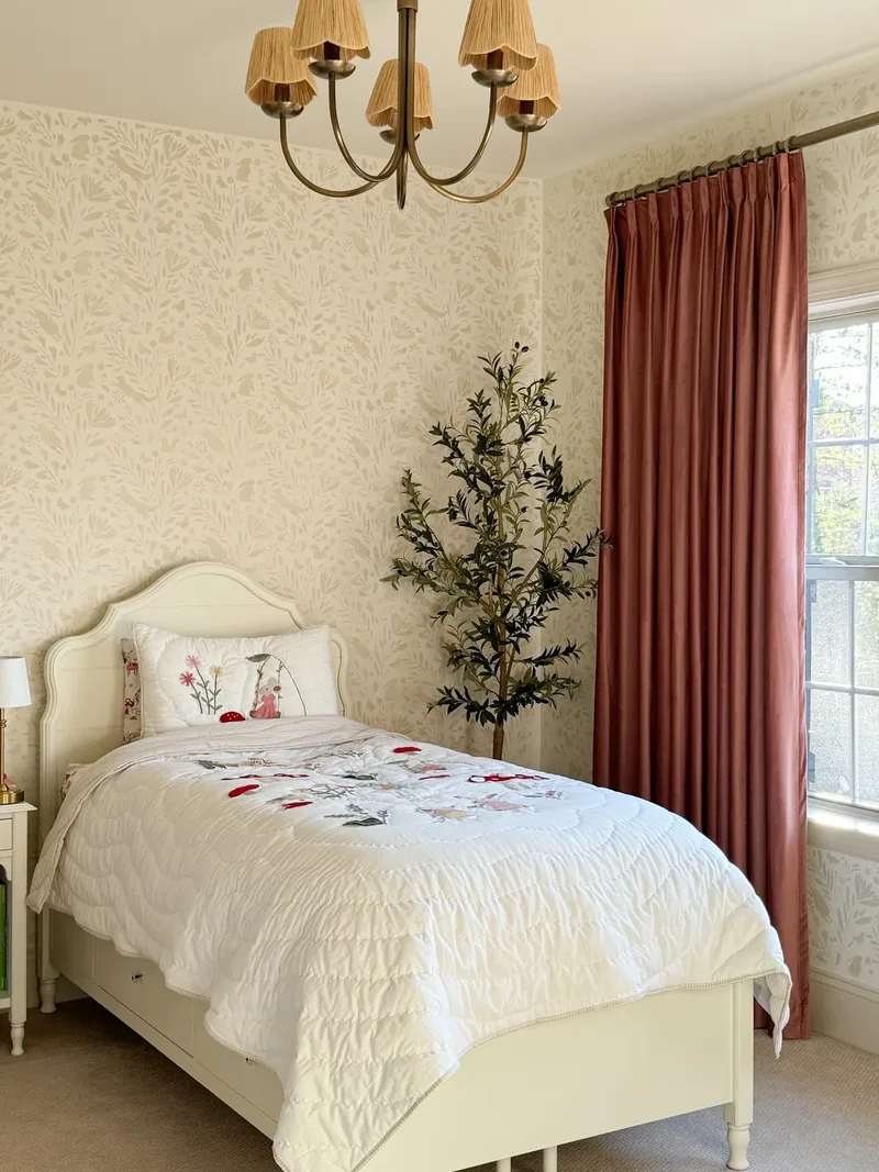 The bed seen lengthwise from the corner, embroidered sham, olive tree, chandelier and rust curtain framing it