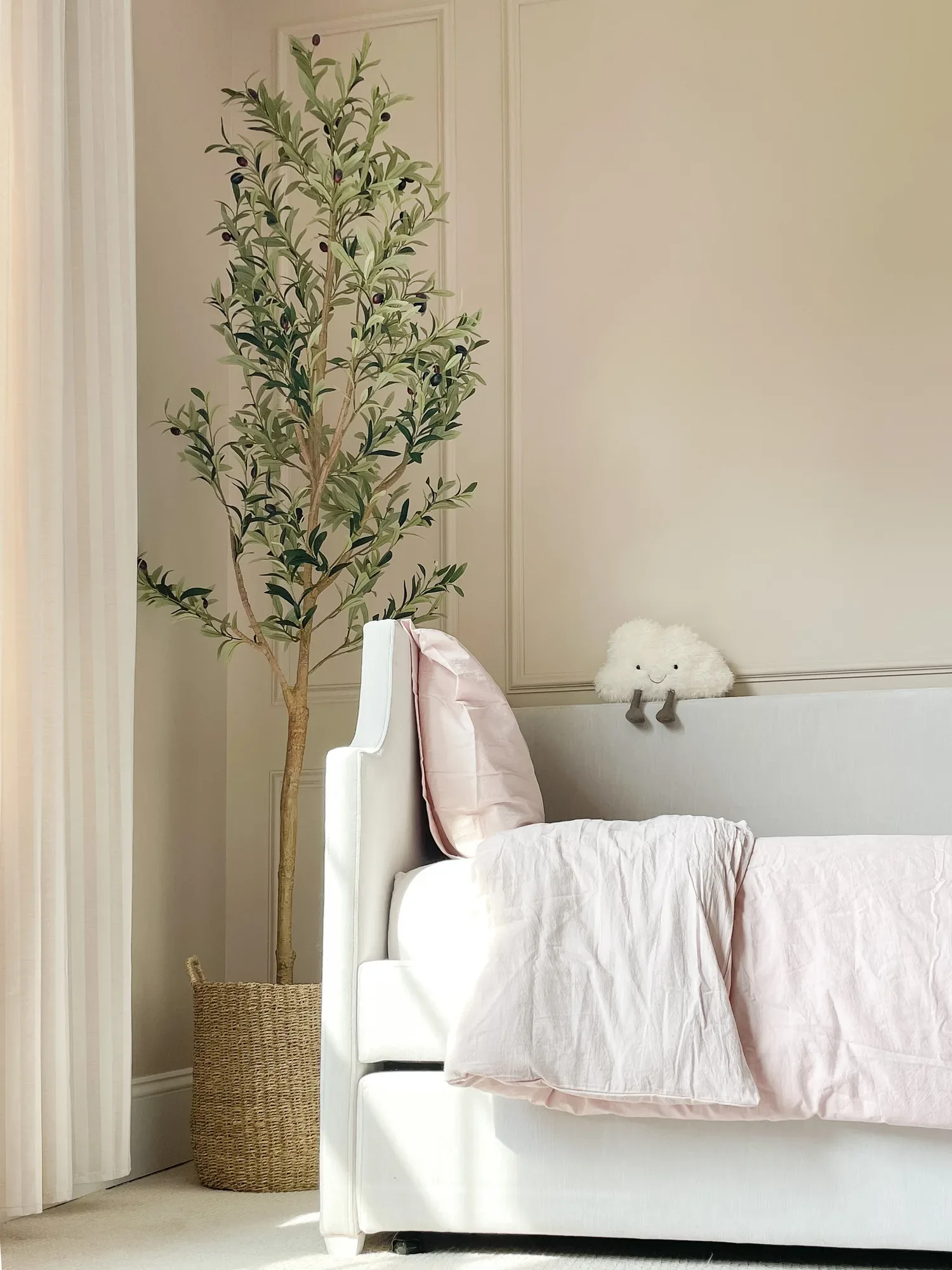 Blush bedding on a white upholstered bed, a plush cloud toy perched on the headboard beside a tall olive tree