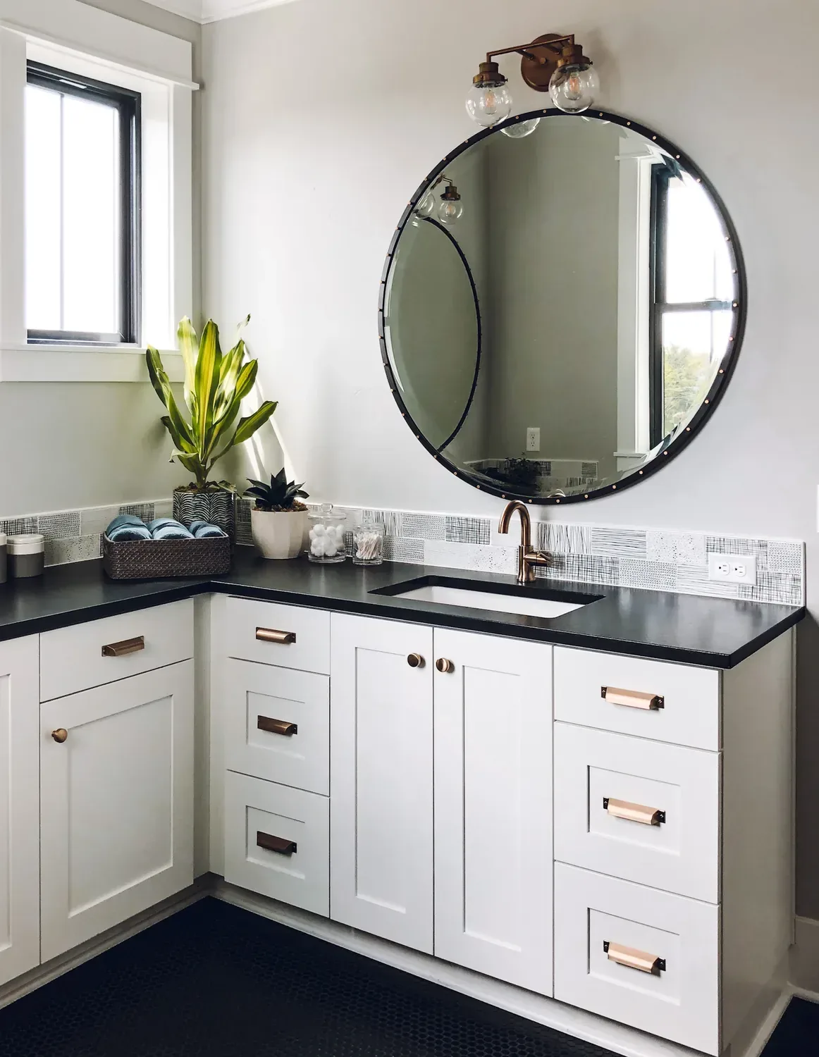 A round black-framed mirror over a white vanity with a black counter and copper handles