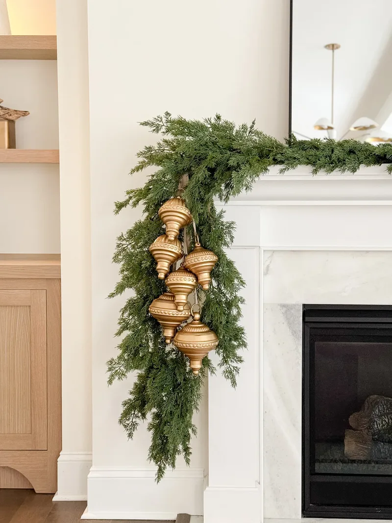 Gold finial ornaments clustered in cedar garland cascading down the corner of a white marble mantel