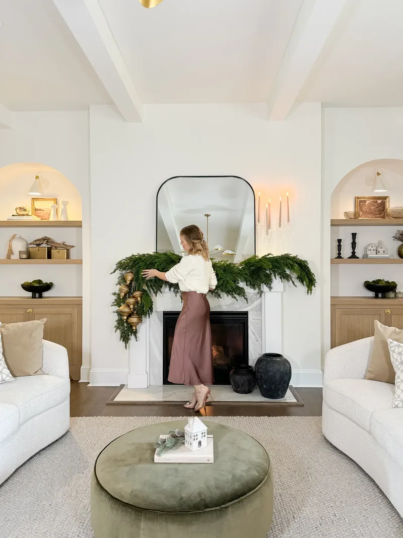 Tatyana arranging cedar garland along the marble mantel between two lit arched built-ins, olive velvet ottoman in front