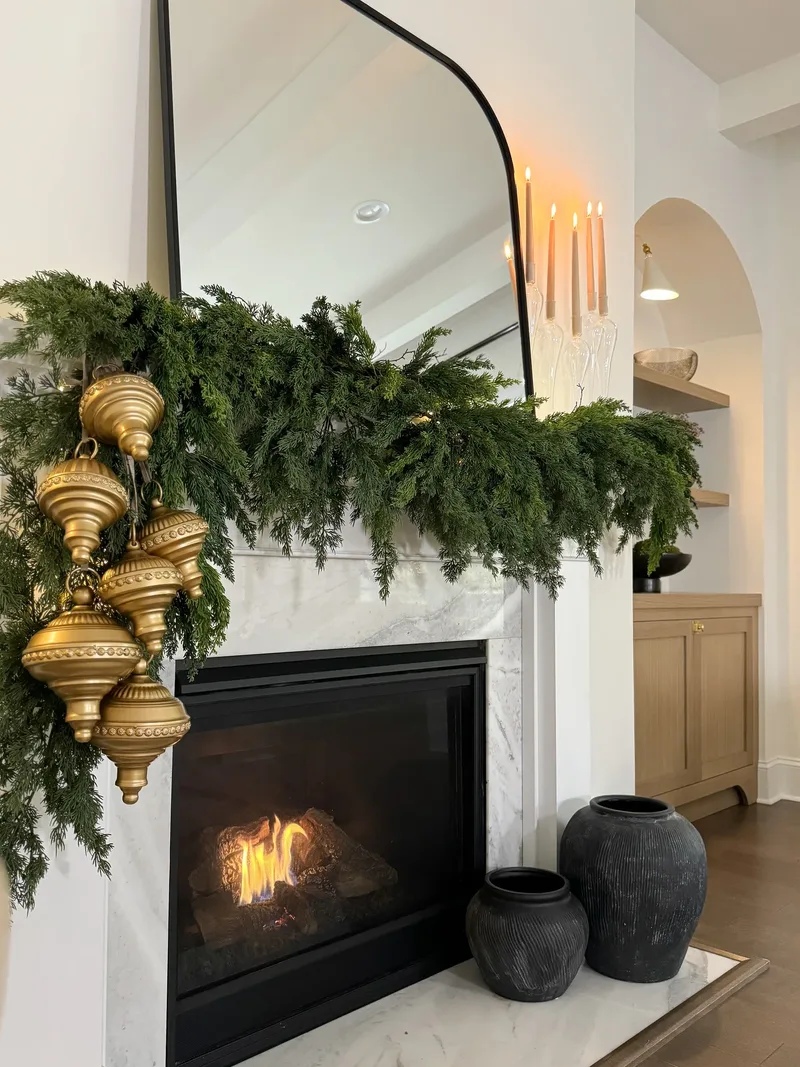Cedar garland and gold finials draped over a lit fireplace, tapers burning beside the arched black mirror
