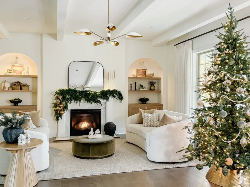 Wide view of the living room: garlanded fireplace, curved cream sofa, tall lit tree at the window