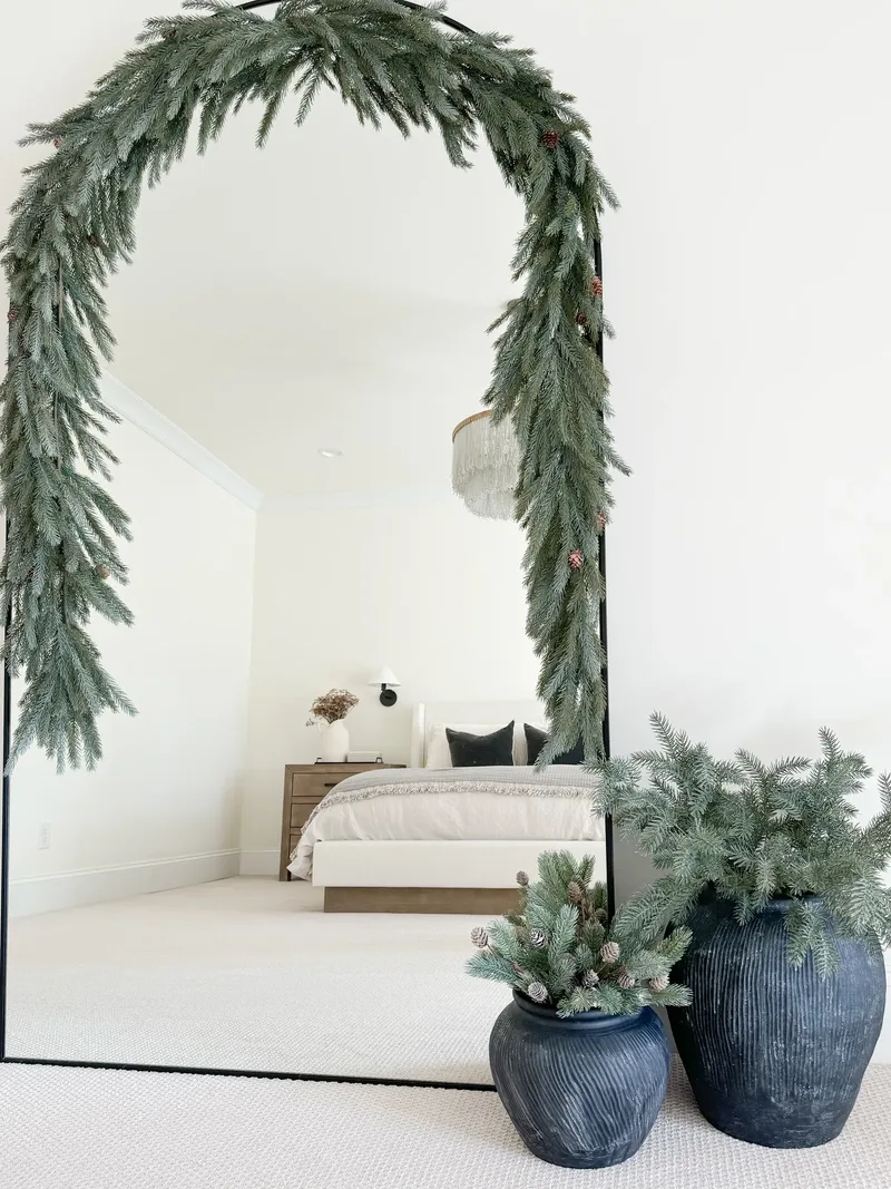 Blue spruce garland draped over an arched floor mirror in a bedroom, black pots of clippings alongside