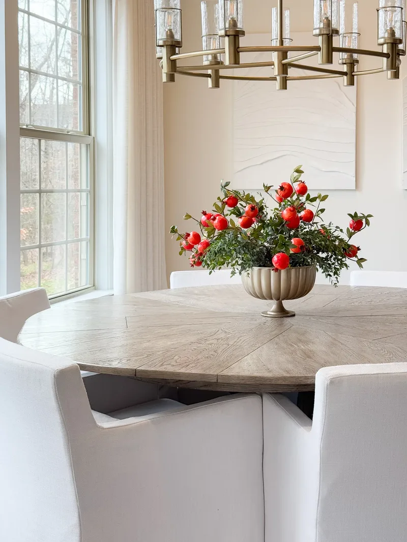Round oak dining table under a brass chandelier, red pomegranate branches in a fluted pedestal bowl