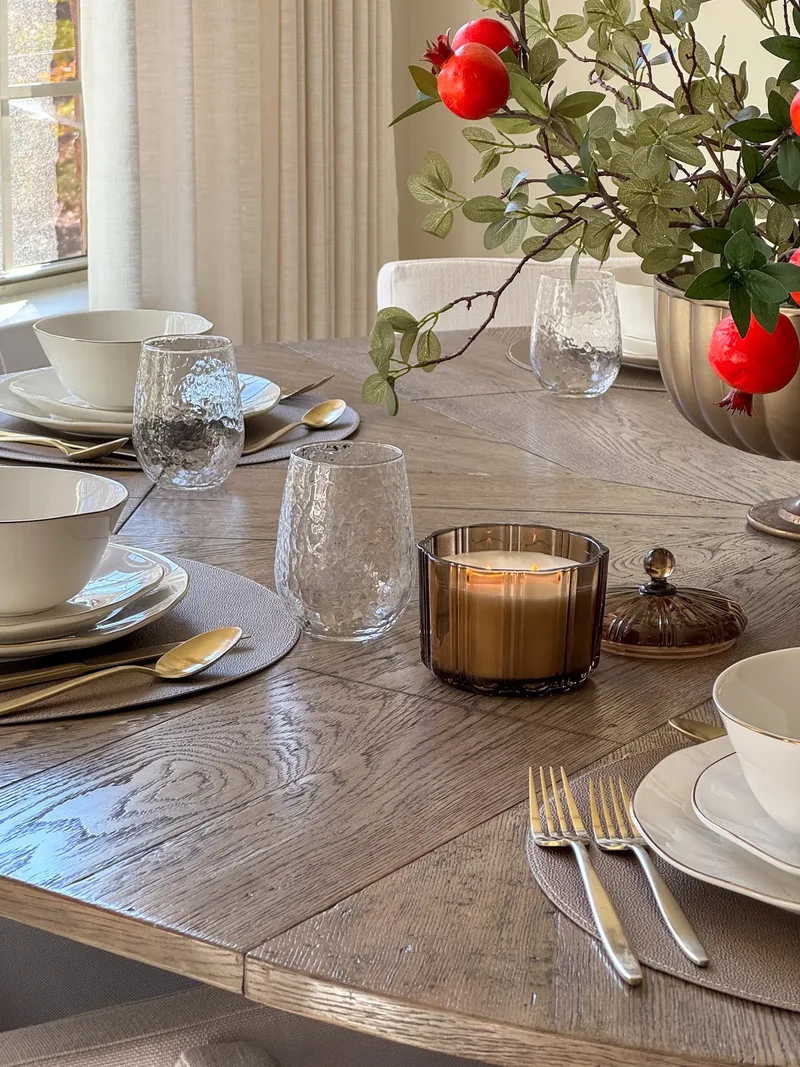 Hammered glass tumblers, gold spoons and a lit amber candle on leather placemats, pomegranate stems behind