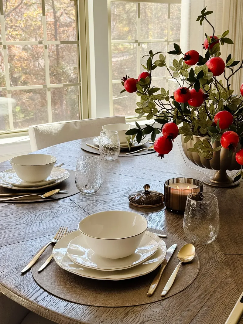 Two settings across the table with pomegranate branches, a lit candle, autumn woods through the windows