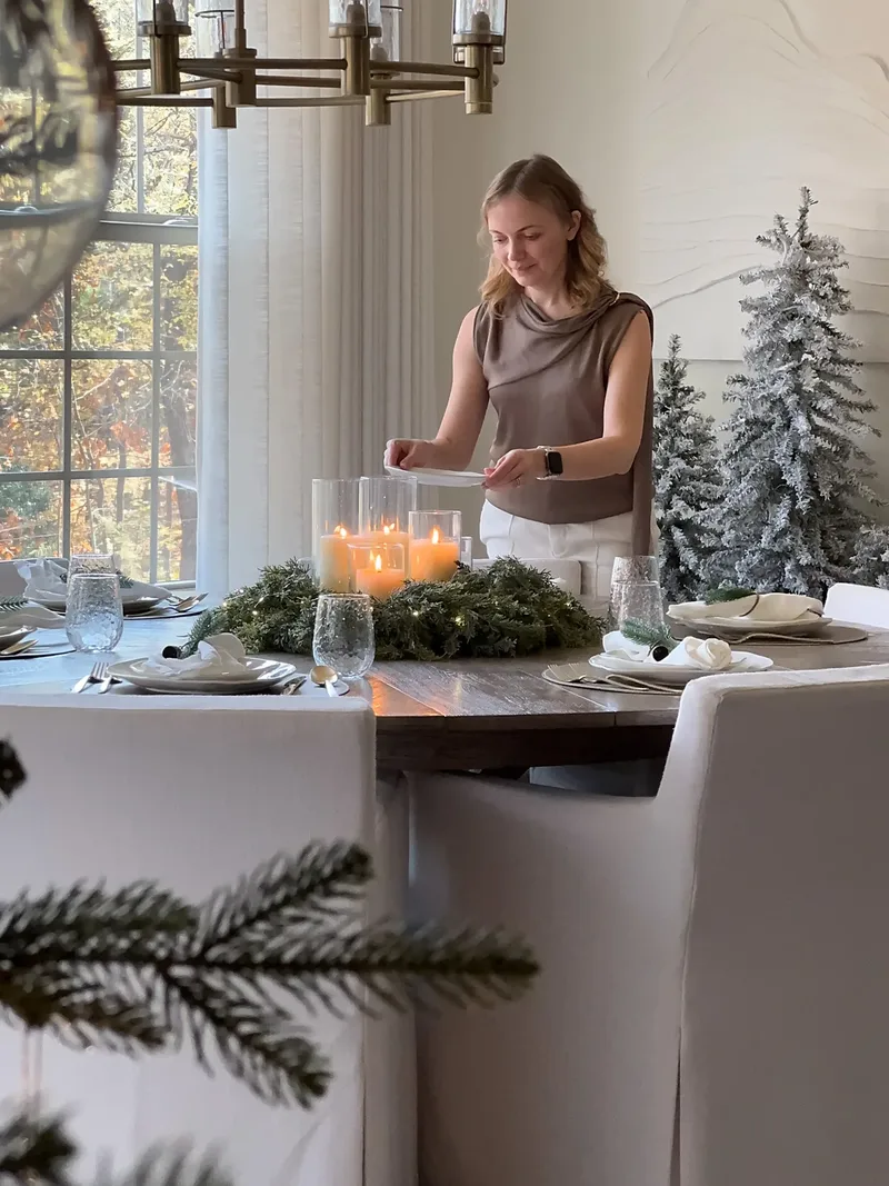 At the dining table, Tatyana holds a long lighter over the candle wreath, flocked trees behind