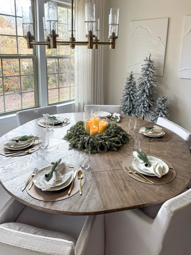 Round table set with pine-sprig napkins and a candlelit fir wreath, flocked trees in the corner