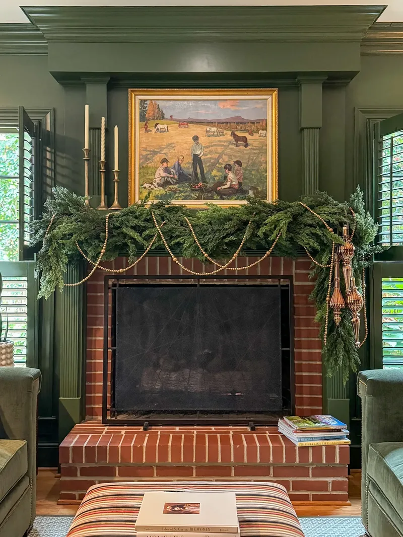 Brick fireplace in a dark green panelled room, cedar garland swagged with wooden beads under a landscape painting