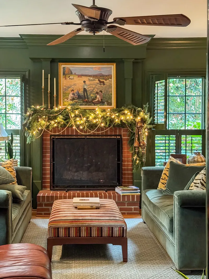 The same green room seen wider with the garland lights on, velvet sofas and a striped ottoman facing the hearth