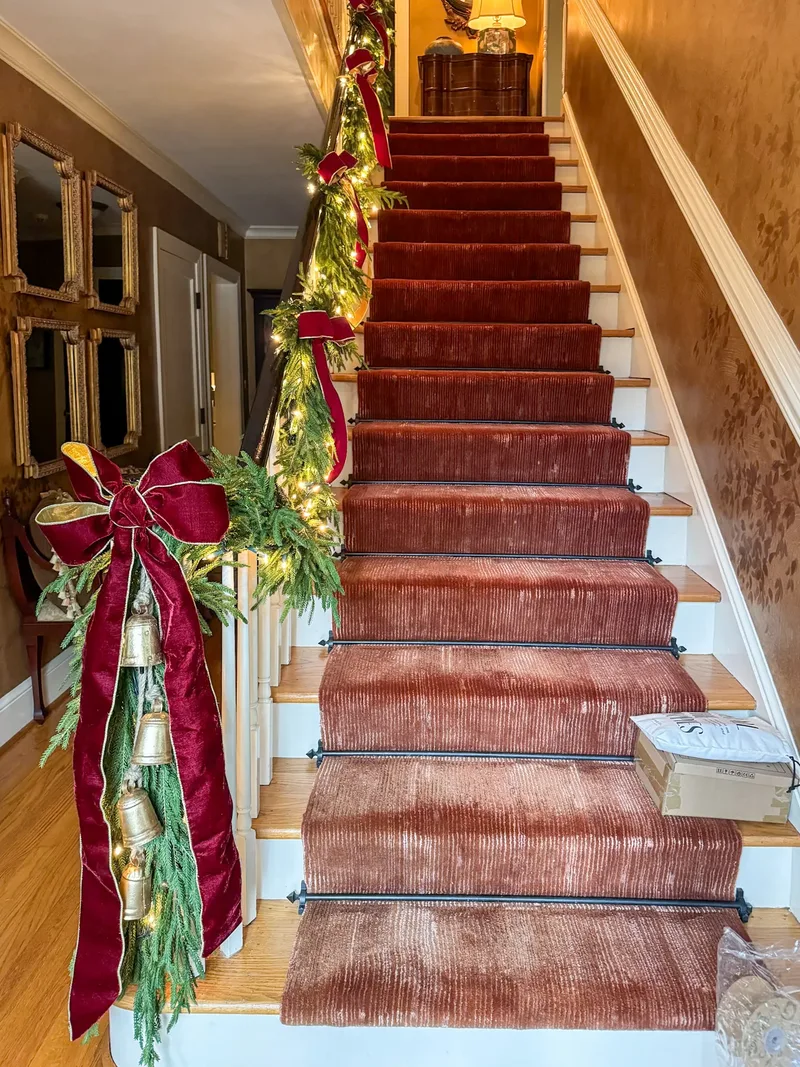 Staircase banister wrapped in lit cedar garland with deep red velvet bows and brass bells, rust velvet runner