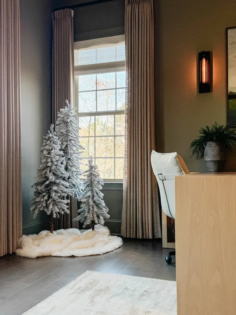 Three flocked pine trees on a faux fur skirt in an office corner beside a curtained window