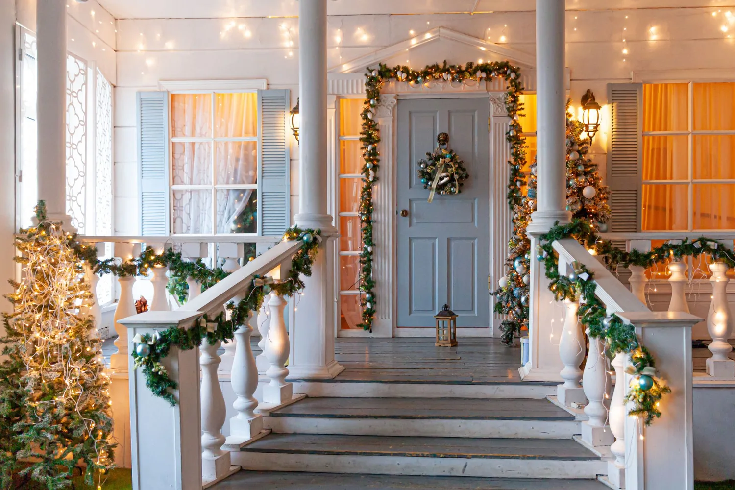 A white porch lit for Christmas, garland along the rails and a wreath on the front door