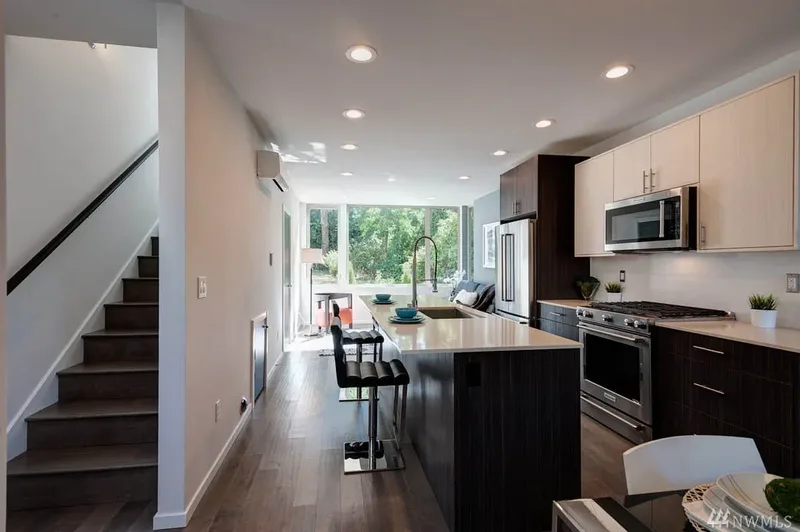 A galley kitchen seen from the stairs, dark-grain lower cabinets and a quartz island with two black stools