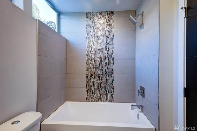 A white alcove tub beneath a shower head, a mosaic tile stripe splitting the grey tiled wall