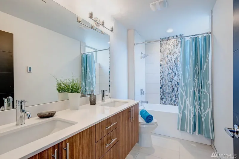 Double-sink vanity in warm wood with a mirror the length of the counter, tub and mosaic tile beyond
