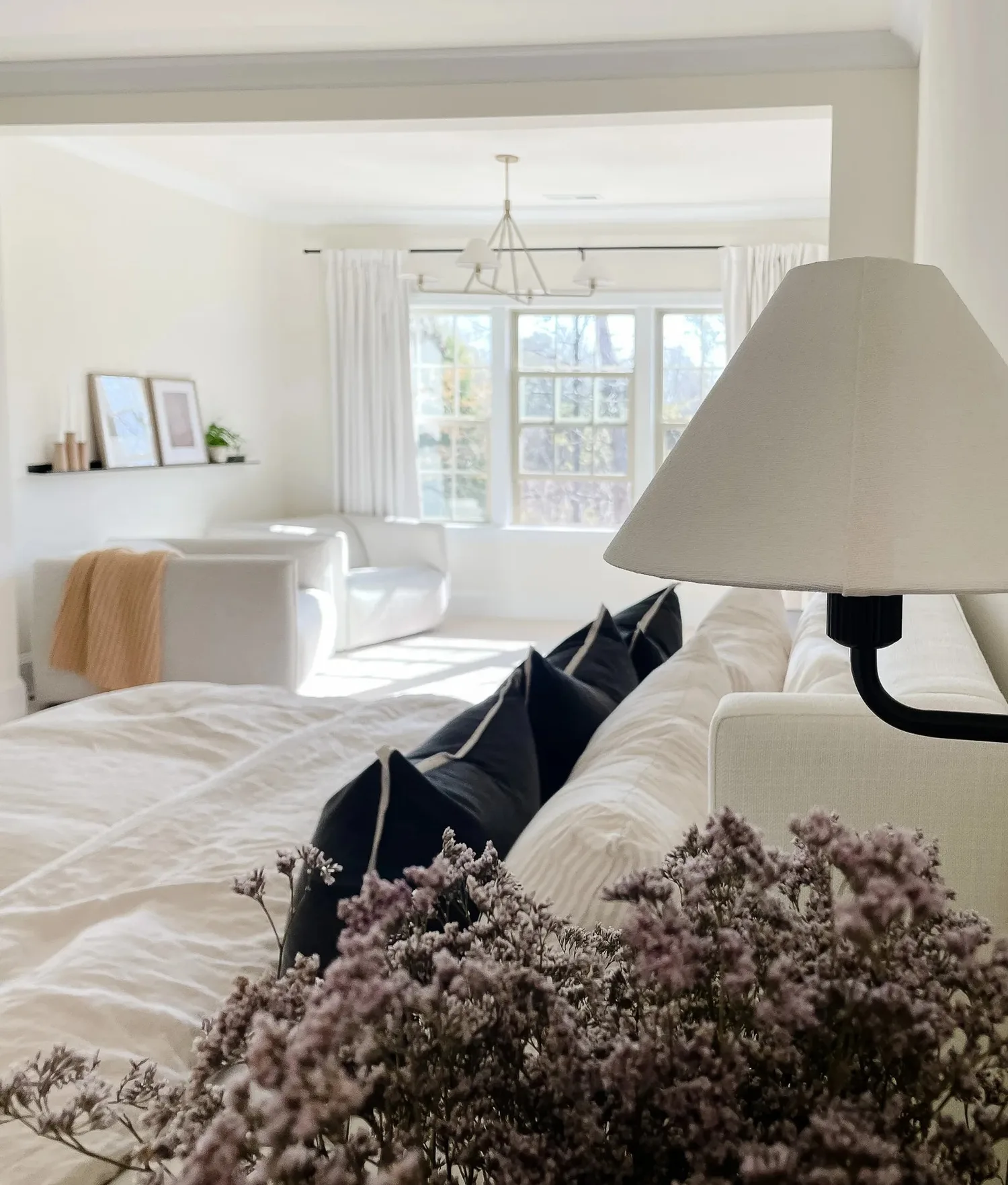 A bright bedroom seen past a linen lamp shade and dried flowers, a sitting area by the windows