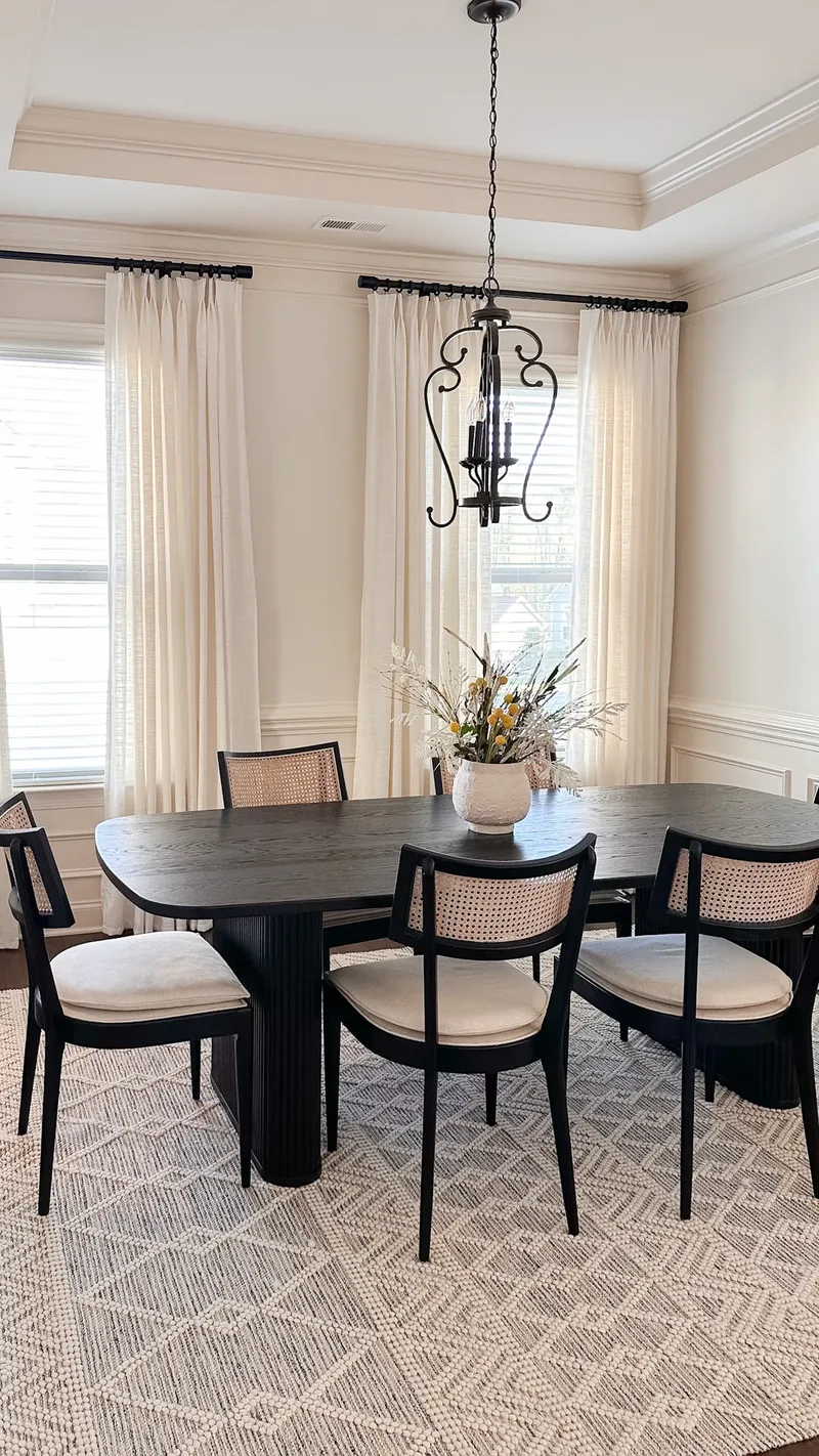 The same black oval table left unset beneath a dark wrought-iron chandelier, dried flowers in a pale vase