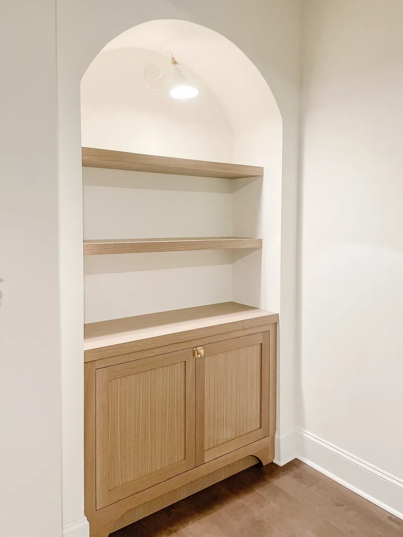 An arched alcove fitted with pale oak shelves and a brass-latched cabinet, still unstyled, lit by a sconce