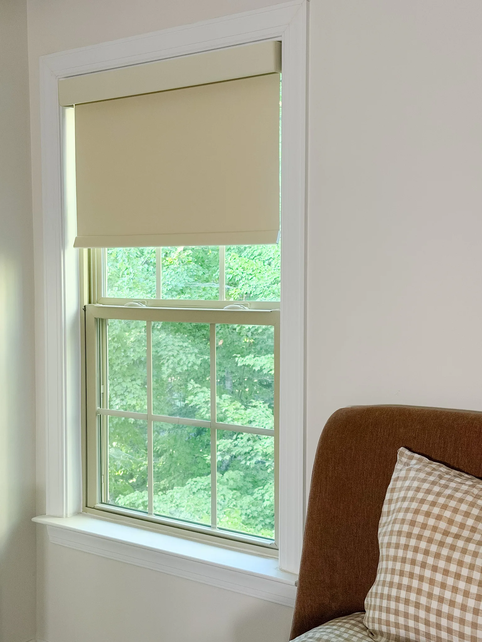 A cream inside-mounted roller shade partially lowered beside a brown bed and gingham pillow