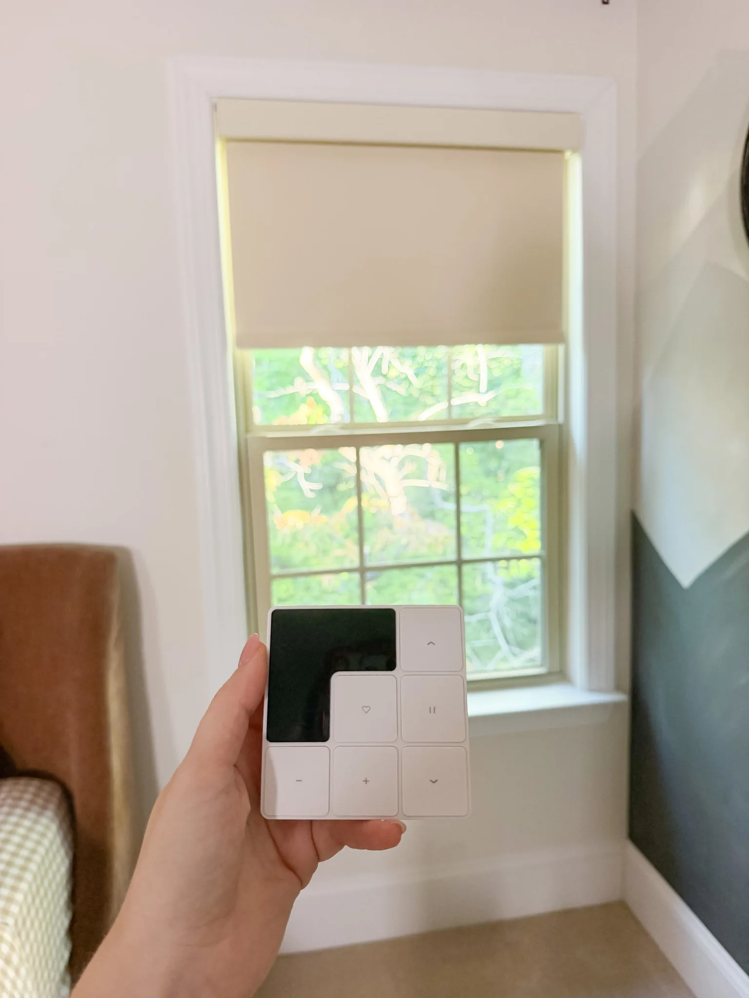 A hand holding the Freshine shade remote in front of a partially lowered bedroom roller shade