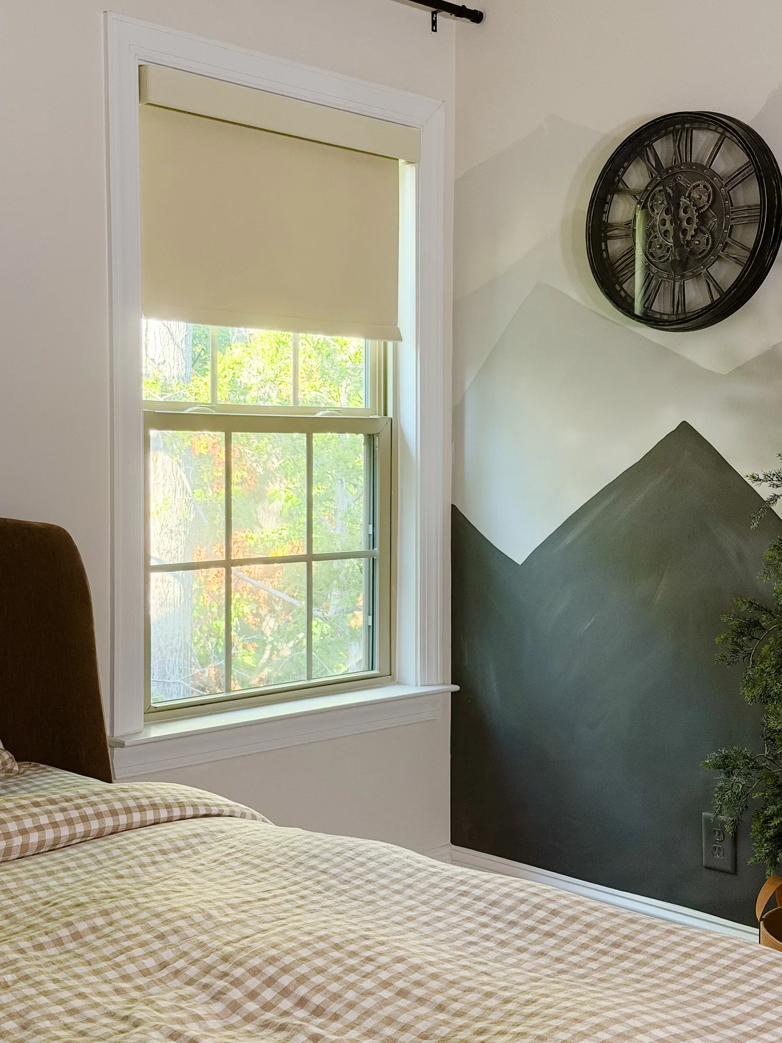 A partially lowered cream roller shade beside a mountain mural and brown upholstered bed
