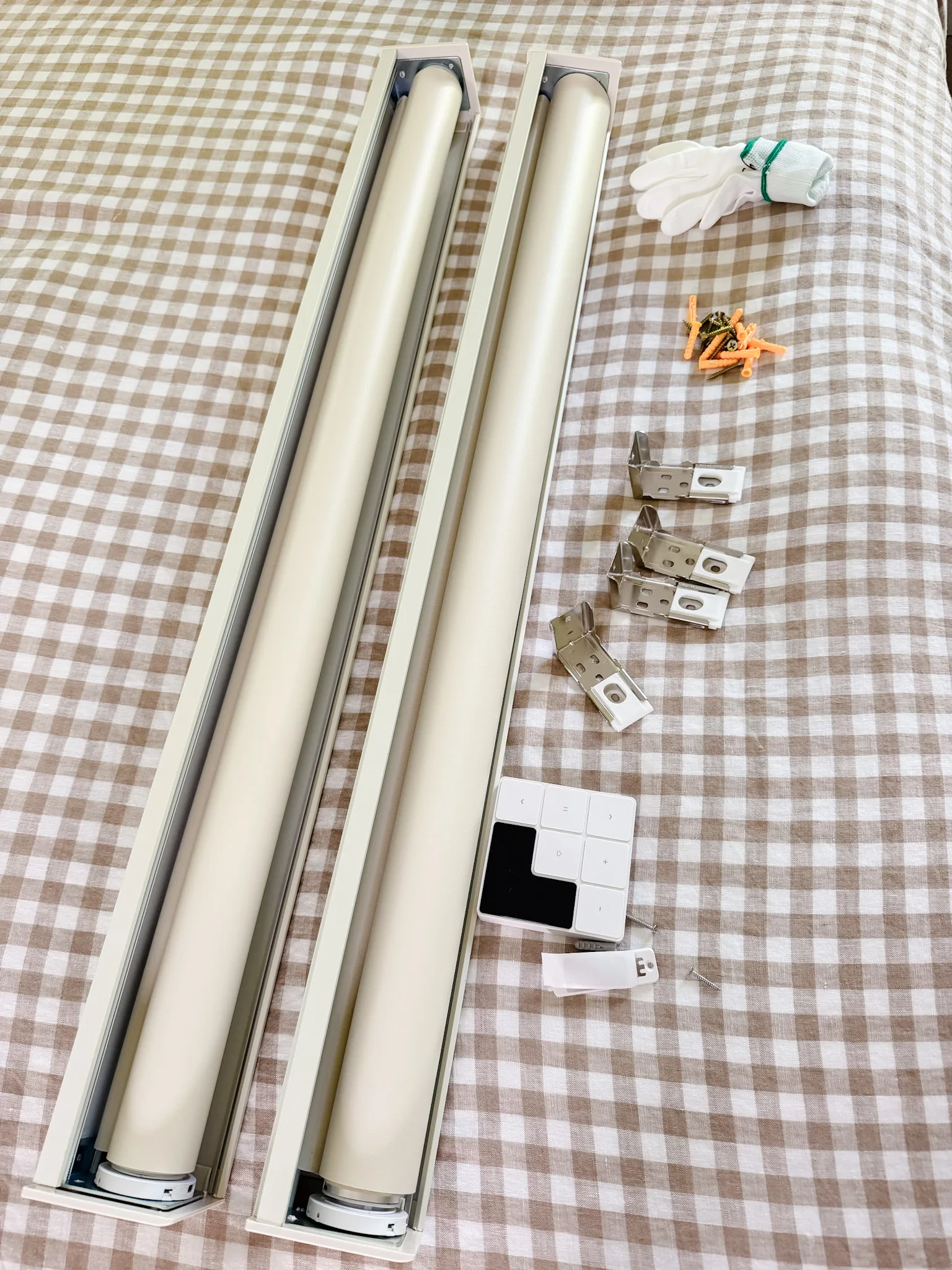 Two cream roller shades with mounting brackets, screws, remote control, and gloves on gingham bedding