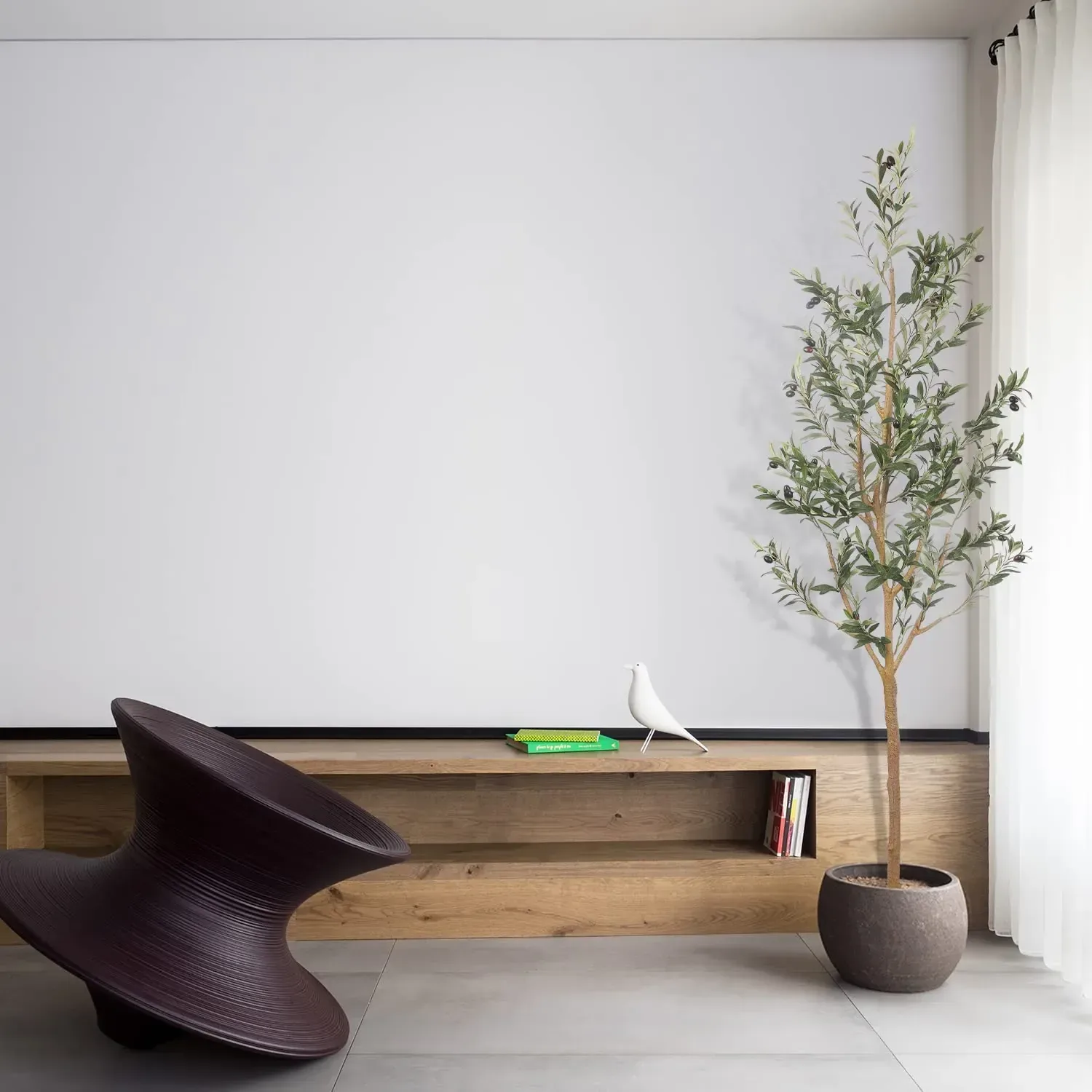 A slim faux olive tree in a stone pot beside a low oak bench and a curved dark chair