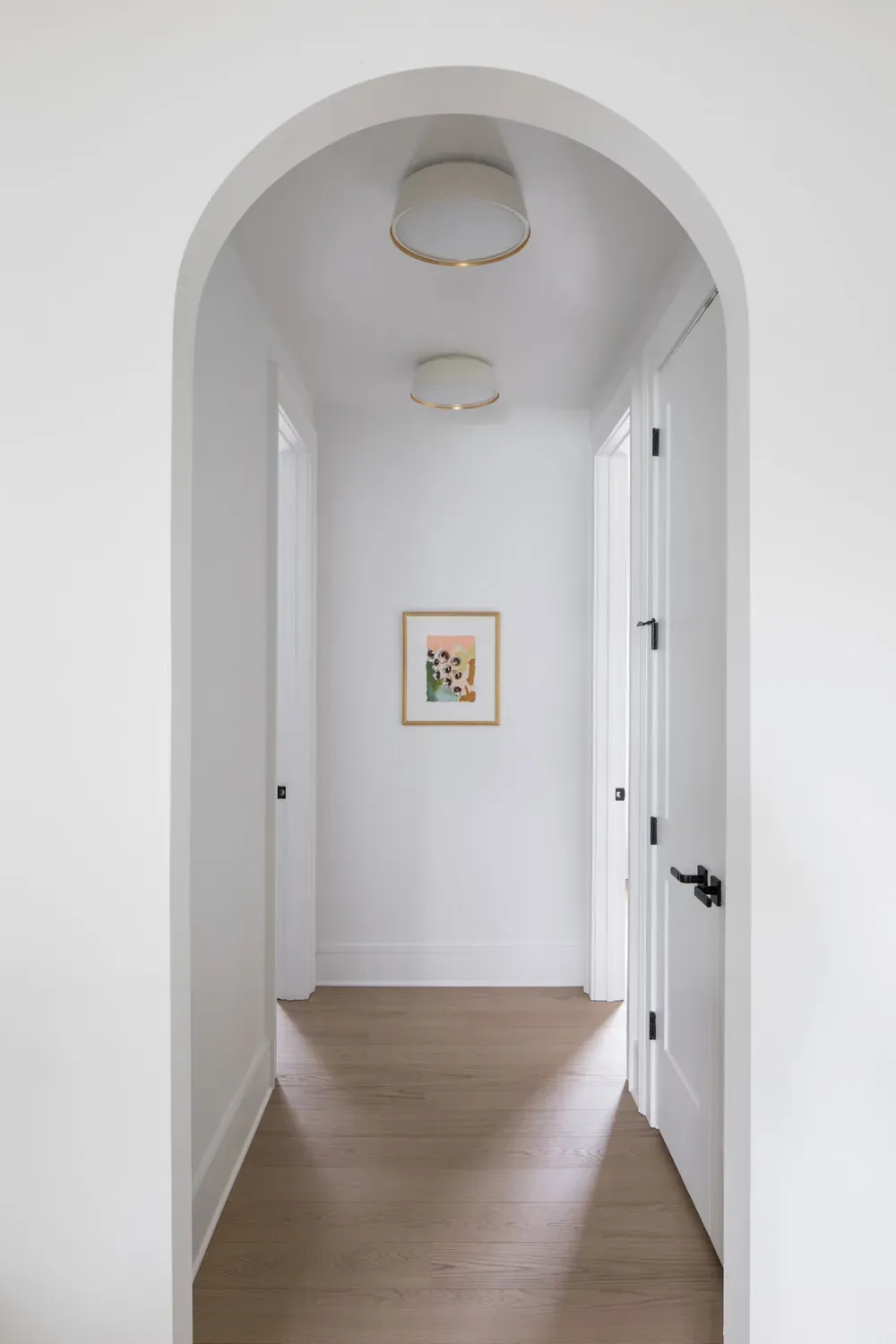 An arched opening onto a white hallway with wood floors and two brass flush ceiling lights