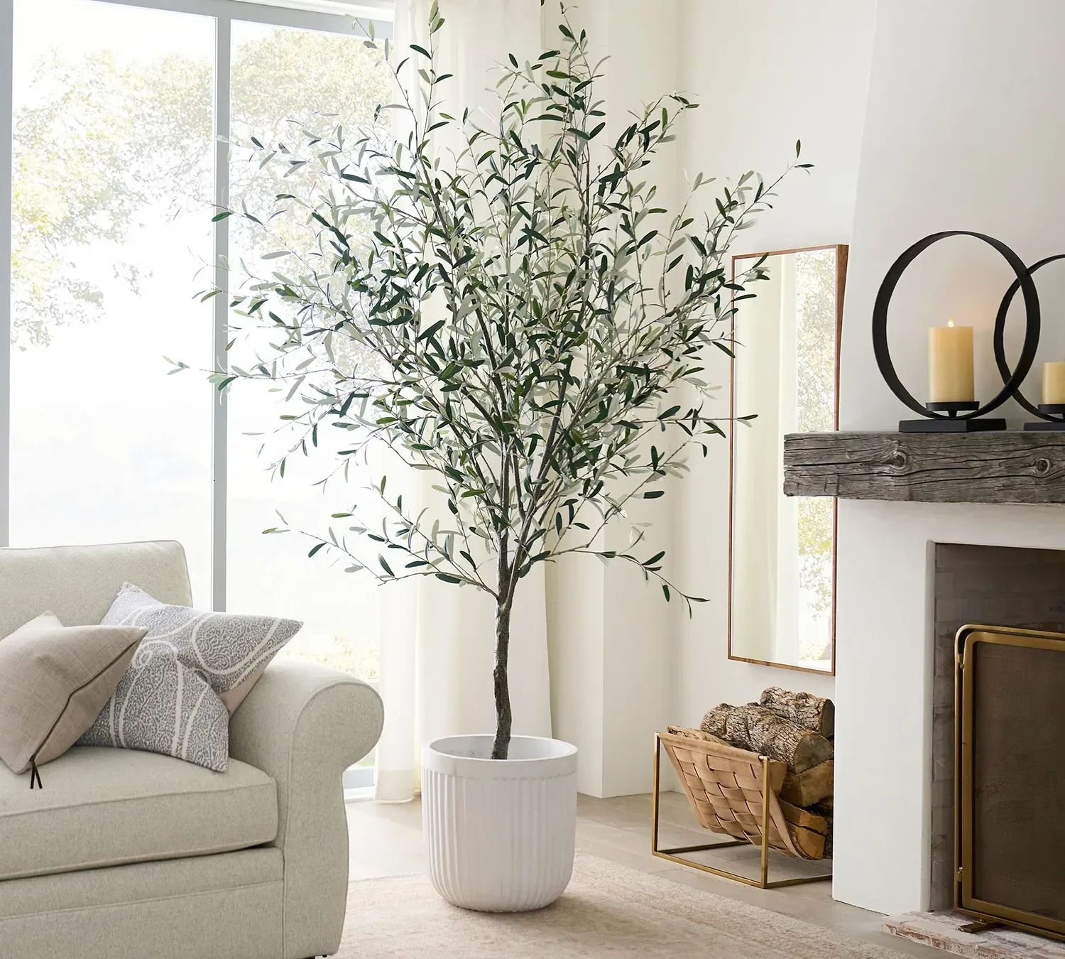 A tall faux olive tree in a white fluted planter beside a fireplace and a cream sofa