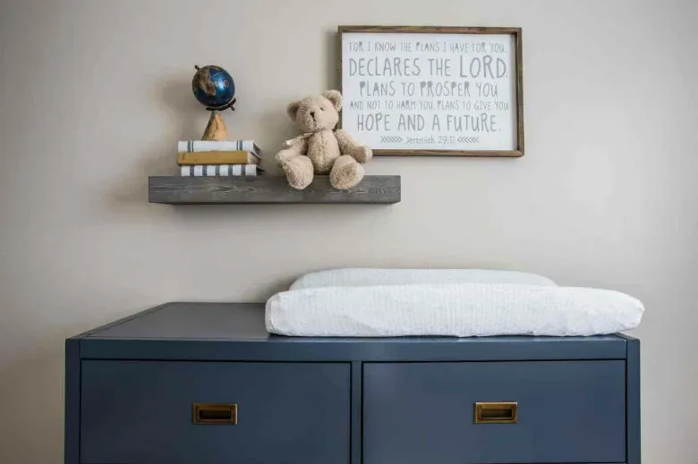 A navy changing dresser below a shelf with a globe, books and a teddy, and a framed scripture