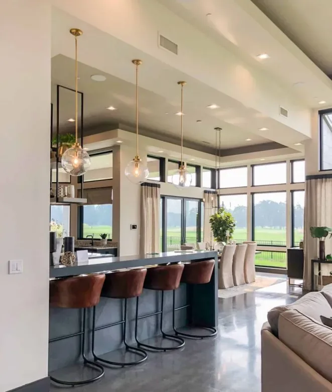 A kitchen island lined with leather bar stools under glass globe pendants, tall windows beyond
