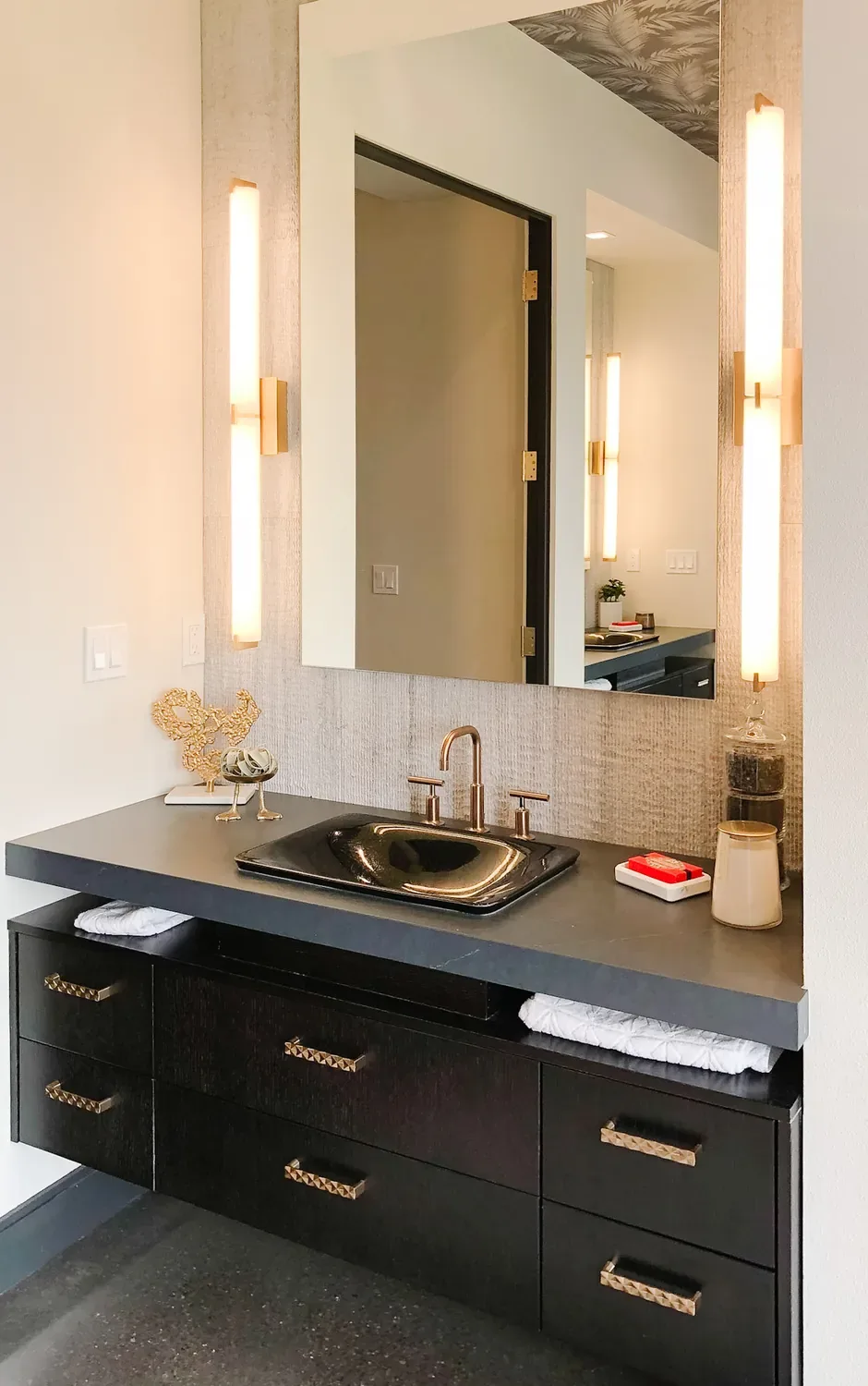 A dark floating vanity with vertical light bars either side of a grasscloth-framed mirror