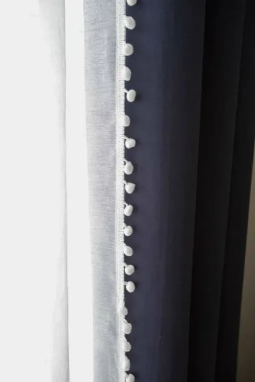 A close view of white pom-pom trim down the edge of the navy curtain panel beside the sheer