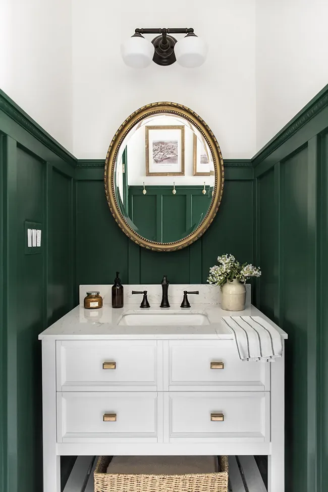A dark green panelled powder room with a gilt oval mirror under a black three-globe light