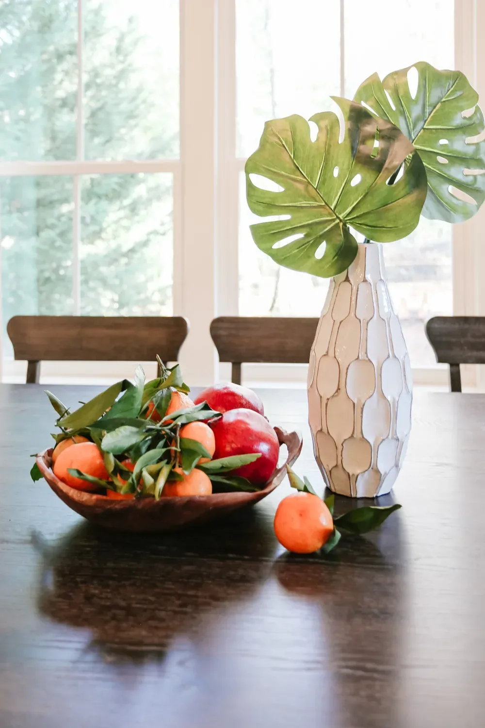 A wooden bowl of clementines and a white honeycomb vase of monstera leaves on the table