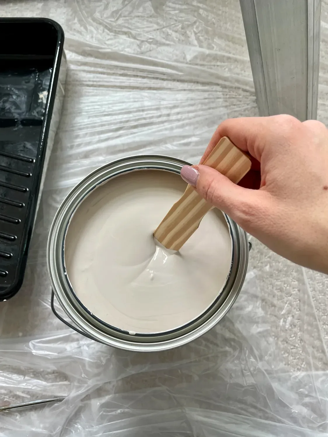 White paint stirred with a wooden stick beside the roller tray on plastic sheeting