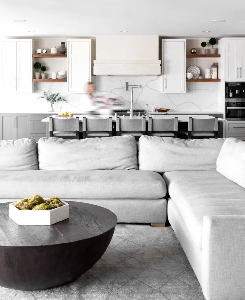 A white kitchen with open shelves and a marble backsplash, seen across a pale sectional from the living room